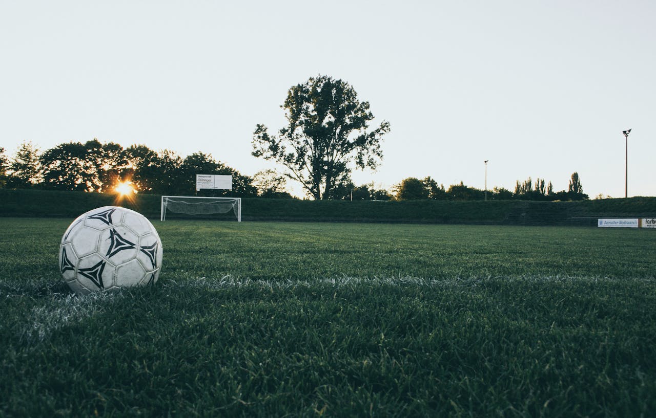 portfolio-02 Soccer ball on field at sunrise, serene landscape with goalpost in background.
