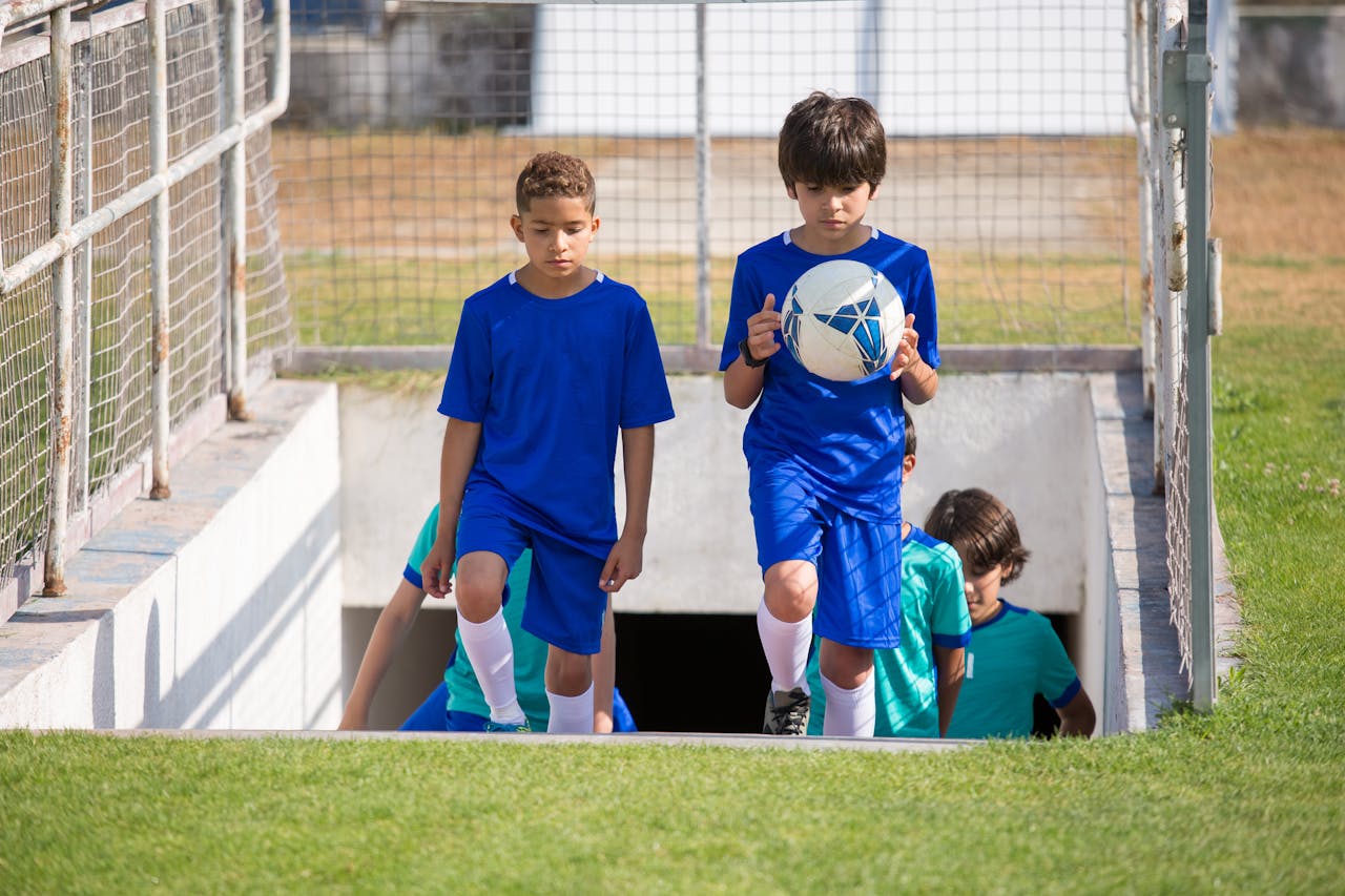 portfolio-05 Children walking onto a soccer field during a sunny day, holding a soccer ball.