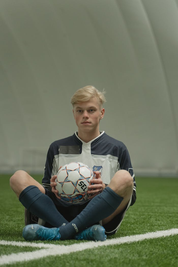 portfolio-01 Teen soccer player in uniform sitting on artificial grass indoors, holding a soccer ball.
